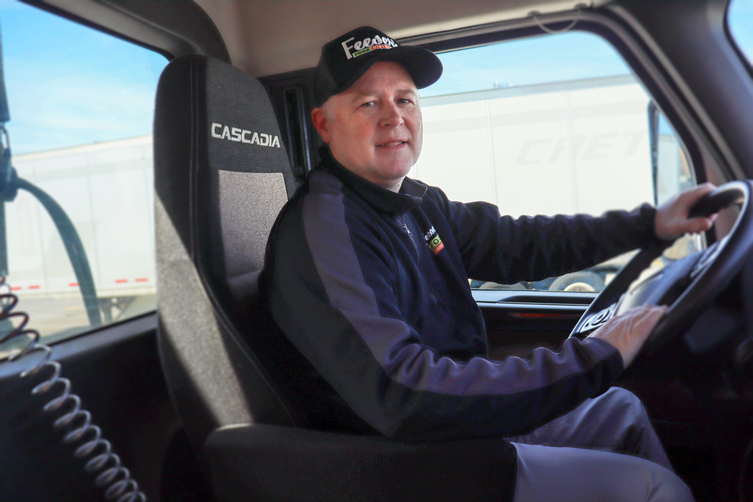 a man smiling and sitting in the driver's seat of a truck wearing a Feeser's quarter zip and baseball cap